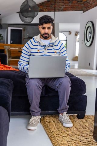 Man working remotely on laptop in modern open-plan loft living room wearing striped hoodie
