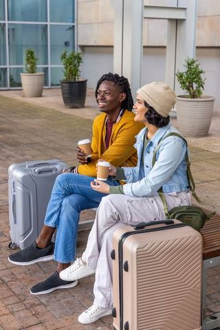 Diverse travelers sipping coffee and smiling on bench with luggage outside modern terminal