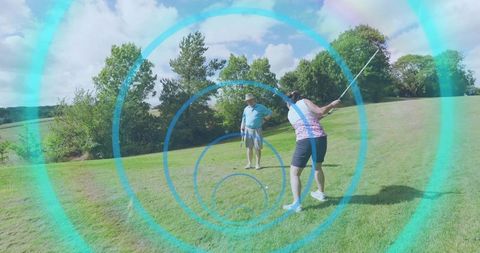 Couple playing golf on sunlit fairway with woman swinging and man watching through turquoise rings