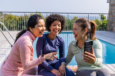 Diverse women in athleisure laughing by pool while sharing smartphone and bonding