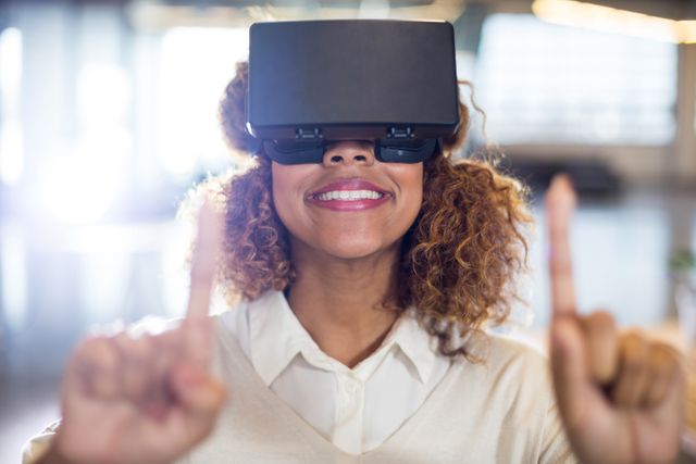 Woman with curly hair smiling while using virtual reality glasses in an office environment. Ideal for illustrating concepts of modern technology, innovation in the workplace, and immersive digital experiences. Suitable for use in articles, blogs, and advertisements related to VR technology, tech advancements, and office productivity.