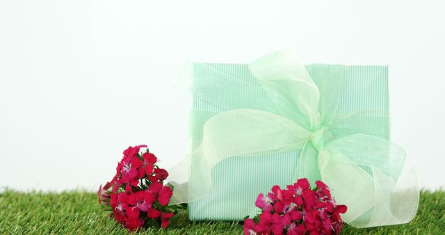 Beautifully wrapped gift box with delicate ribbon placed on green grass, surrounded by vibrant red flowers. Ideal for use in design projects related to celebrations, holidays, or special occasions. Can be used for greeting card designs, promotional material, and website banners.