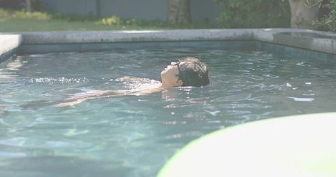 Sunlit backyard pool floating Asian man wearing sunglasses relaxing on back near inflatable