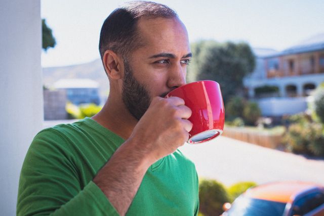 Man enjoying a cup of coffee on a sunny balcony, perfect for themes of relaxation, morning routines, and quarantine lifestyle. Ideal for use in articles or advertisements related to home living, self-care, and leisure activities.