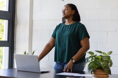 African american woman standing at desk using laptop reviewing papers in bright workspace