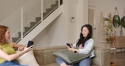 Diverse female friends relaxing on sofa, scrolling smartphones in cozy modern lounge