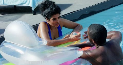 Diverse couple clinking drinks on rainbow inflatable, relaxing by pool edge, summer vibes