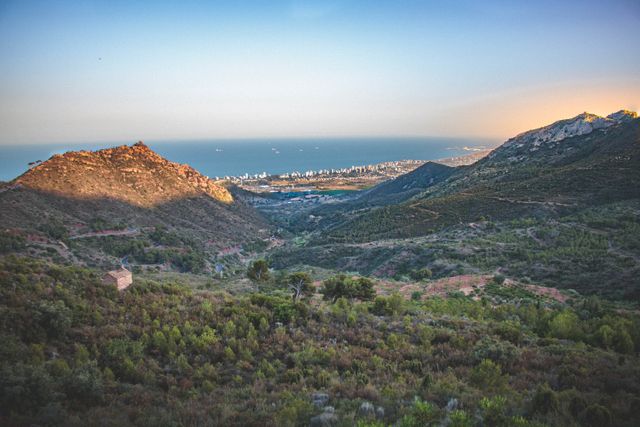 Scenic aerial view of rugged mountains leading down to a coastal city at sunset. Green hills with patches of trees contrast with the blue sky and distant ocean. Ideal for use in travel brochures, nature-inspired publications, or advertising outdoor activities and adventures.