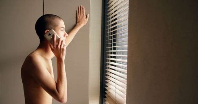Young man talking on the phone while gazing out of a window with blinds from indoors. Perfect for concepts such as communication, technology, introspection, casual behavior, and everyday life moments.