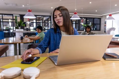 Young asian woman working on laptop in modern coworking space reaching for smartphone