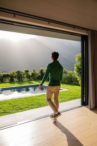 Man standing watching sunlight streaming through sliding glass door overlooking pool and mountains