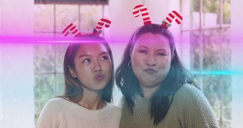 Playful holiday duo puckering faces wearing candy cane headbands in sunlit window