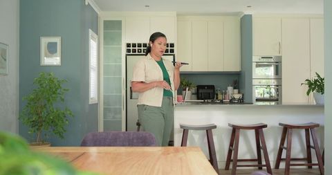 Asian woman speaking into smartphone using wireless earbud in modern open-plan kitchen