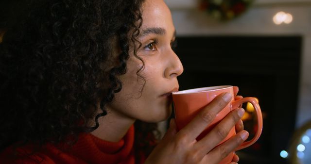 Young woman enjoying hot beverage indoors with curly hair, drinking from orange mug, suggests relaxation and comfort. Ideal for content about lifestyle, self-care, relaxation indoor activities, winter months and warm beverages.