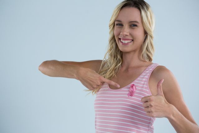 This image shows a smiling woman pointing at a pink ribbon on her shirt while giving a thumbs up. It is perfect for promoting breast cancer awareness, health campaigns, charity events, and educational materials about cancer prevention and support.