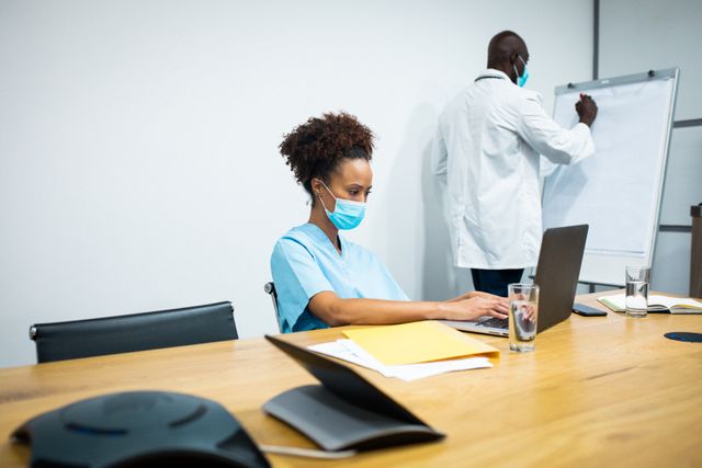 Diverse female and male doctor wearing face masks using laptop and writing on display pad in office. medical and healthcare services during coronavirus covid 19 pandemic.