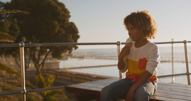 Curly-haired child sitting on bench at seaside pier, enjoying an ice cream cone during sunset. Background includes blurred elements of trees, water, and piers. Suitable for use in advertisements about family vacations, summer activities, childhood moments, and leisure time. Fits themes of outdoor fun, relaxation by the sea, and tasty treats.