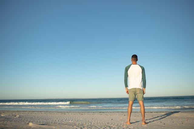 Man standing on sandy beach facing the ocean under a clear sky at sunset. Ideal for travel, vacation, relaxation, or outdoor lifestyle themes. Perfect for promoting beach holidays, mental wellness, and serene getaways.