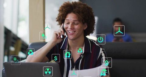 Smiling man talking on phone while working at laptop with renewable energy icons overlay