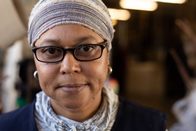 Portrait of a happy biracial woman wearing reading glasses and headscarf and looking at camera in the workshop at a hat factory. 