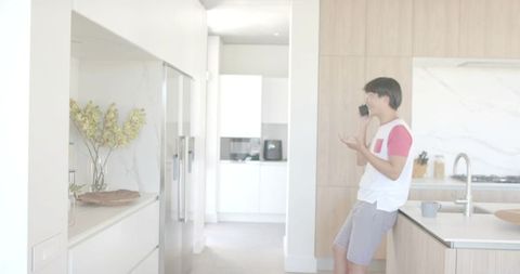 Asian man leaning on kitchen island talking on smartphone in modern open-plan kitchen