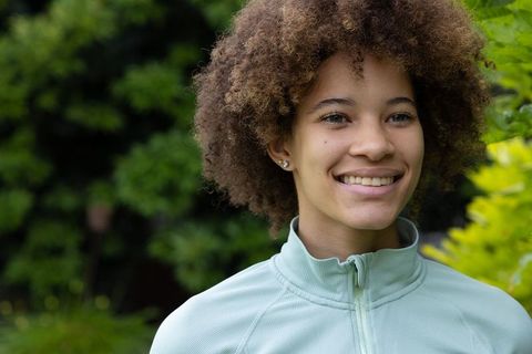 Curly-haired woman smiling in lush garden wearing mint zip-up jacket