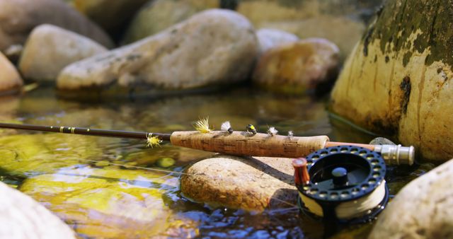 Fly fishing rod and reel are resting on a bed of smooth river rocks in a shallow stream. A few fishing flies are attached to the rod handle. Ideal for promoting fishing gear, outdoor hobbies, nature-themed content, and leisure activities. Perfect for blogs, advertisements, and articles about fishing tips and adventures.