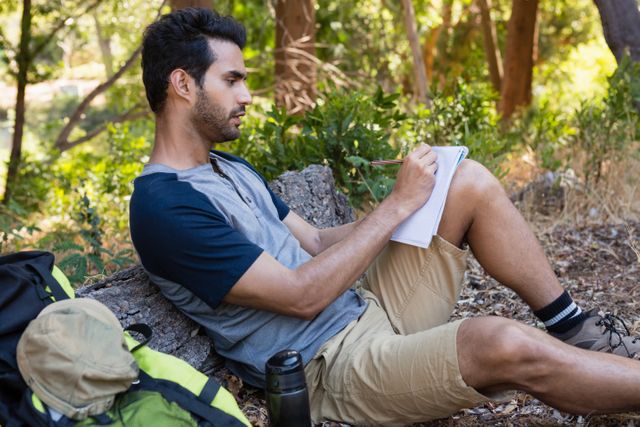 Man writing on notepad while resting on tree trunk in the forest