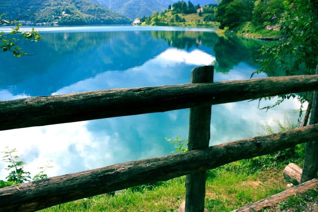 Tranquil lake with clear water reflecting blue sky and mountains viewed through wooden fence. Ideal for travel blogs, nature magazines, and landscape backgrounds.