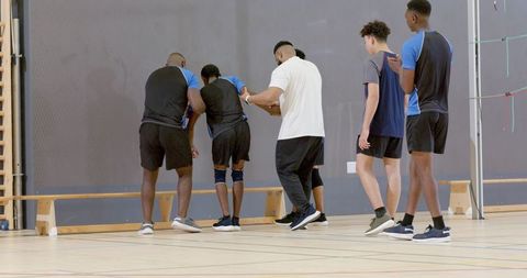 Diverse male athletes and coach supporting hurt teammate in indoor court training drill