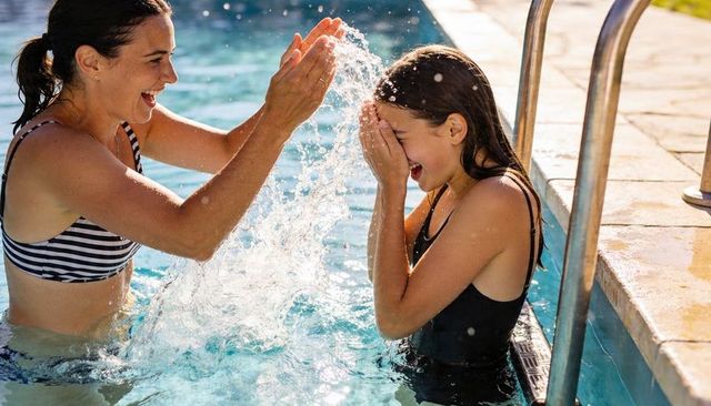Woman splashing girl laughing in sunlit outdoor pool for summer family water play