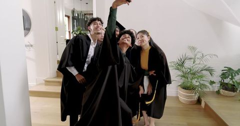Graduate Friends Taking Selfie at Home Wearing Caps and Gowns Celebrating Achievement