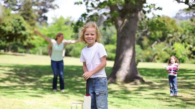 Young boy holding a cricket bat in green park, family engaged in game activity in background. Ideal for promoting family outings, outdoor activities, childhood fun, cricket context, or summer adventures.