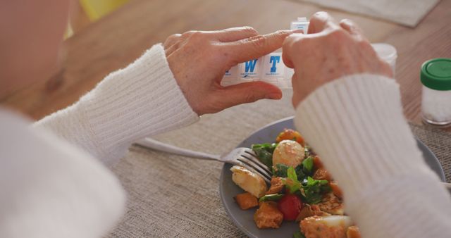 Midsection of senior caucasian woman having lunch at kitchen table opening pill organiser. Medicine and healthcare, illness, mental health, domestic life and senior lifestyle, unaltered.