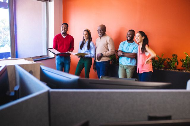 Group of diverse business colleagues standing and smiling while discussing work against an orange wall in a modern office. Perfect for illustrating teamwork, workplace diversity, professional collaboration, and corporate settings in business and marketing materials.