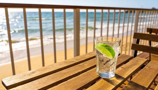 Sparkling lime water on sunlit wooden deck overlooking ocean waves and beach refreshing