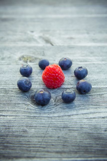 Fresh raspberry encircled by blueberries on weathered wooden surface. Perfect for promoting healthy eating, summer recipes, organic products, nutrition articles, or farmhouse-style decor. Bright red raspberry contrasts with dark blueberries and rustic background emphasizes natural feel.
