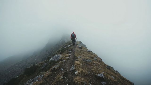 Solo hiker trekking narrow foggy mountain ridge in red jacket, moody alpine solitude