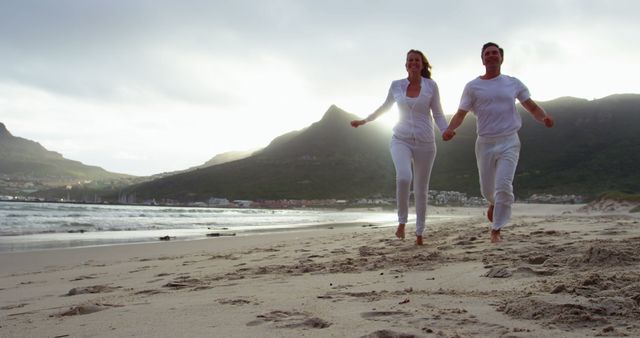 A young Caucasian couple is joyfully running along a sandy beach at dusk, with copy space. Their relaxed white attire and the serene coastal landscape suggest a romantic getaway or a leisurely vacation moment.