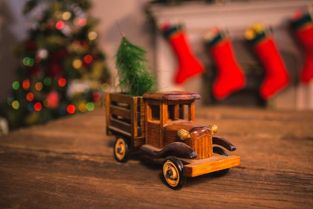 Vintage toy truck carrying a small Christmas tree on a wooden table with Christmas stockings and lights in the background. Perfect for holiday-themed advertisements, nostalgic Christmas cards, and festive home decor inspiration.