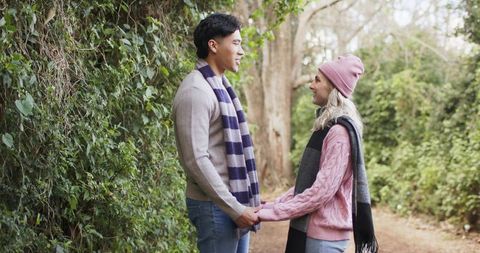 Young couple holding hands on forest trail smiling in knit scarves and beanie, cozy outdoor moment
