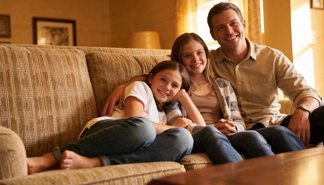 Father hugging two daughters on beige sofa smiling in sunlit living room