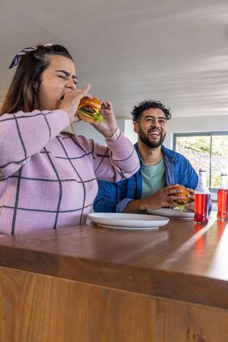 Young couple enjoying cheeseburgers at bright modern kitchen counter laughing with sodas