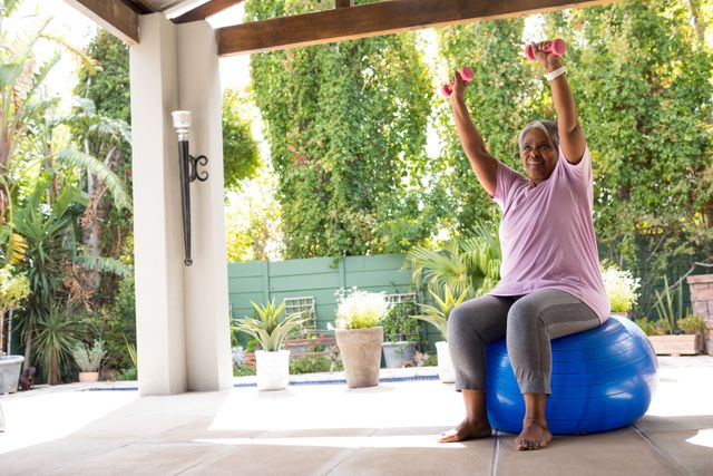 Woman with arms raised holding dumbbells while sitting on fitness ball