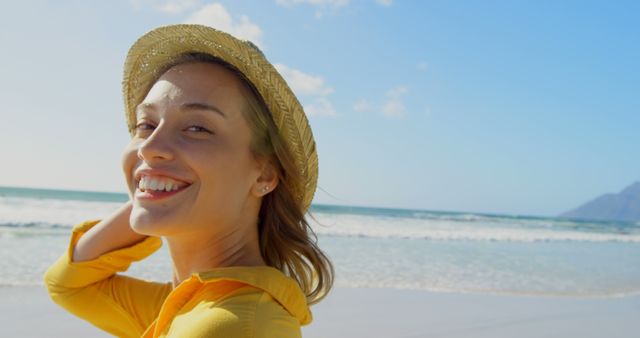 A young Caucasian woman smiles brightly, wearing a straw hat on a sunny beach, with copy space. Her cheerful expression and the coastal backdrop suggest a relaxed vacation atmosphere.
