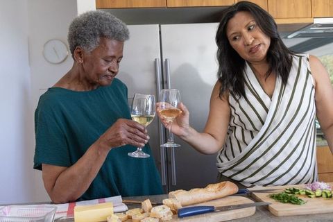 Senior african american and asian women toasting with wine preparing bread and cheese