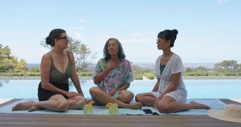 Three women relaxing and chatting by infinity pool on sunny terrace with cocktails