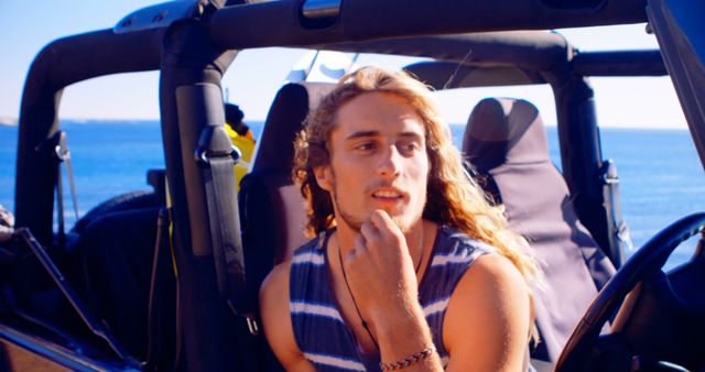 Young man with long hair sitting in a convertible, parked near the ocean. He is looking thoughtful, wearing a sleeveless striped shirt. Background includes clear ocean water and blue sky, suggesting a tropical beach. Ideal for use in advertisements, travel magazines, and lifestyle blogs promoting beach vacations, summer activities, and car rentals.