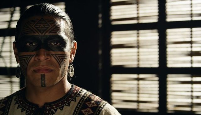 Man wearing geometric face paint and metal hoops, posing in embroidered top under blinds