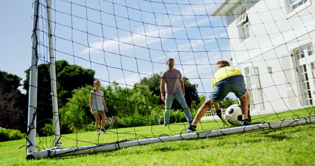 This image captures a family's playful moment playing soccer in the backyard. The father is watching as the two children energetically engage in the game, with one child poised as the goalkeeper, ready to stop the ball. Beautiful weather, a sunny day and lush green garden setup the perfect backdrop for family bonding and outdoor recreation. Ideal for use in advertisements promoting family activities, outdoor sports, recreational equipment, or parenting resources.